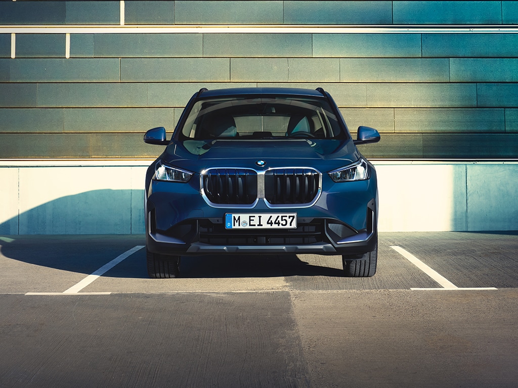 BMW X1 (U11) - In einem Parkhaus, mit einer Wand, die das Sonnenlicht im Hintergrund reflektiert. Die angrenzenden Parkplätze sind leer.