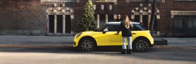 MINI Junge Gebrauchte, gelber MINI mit Kundenübergabe vor urbaner Kulisse