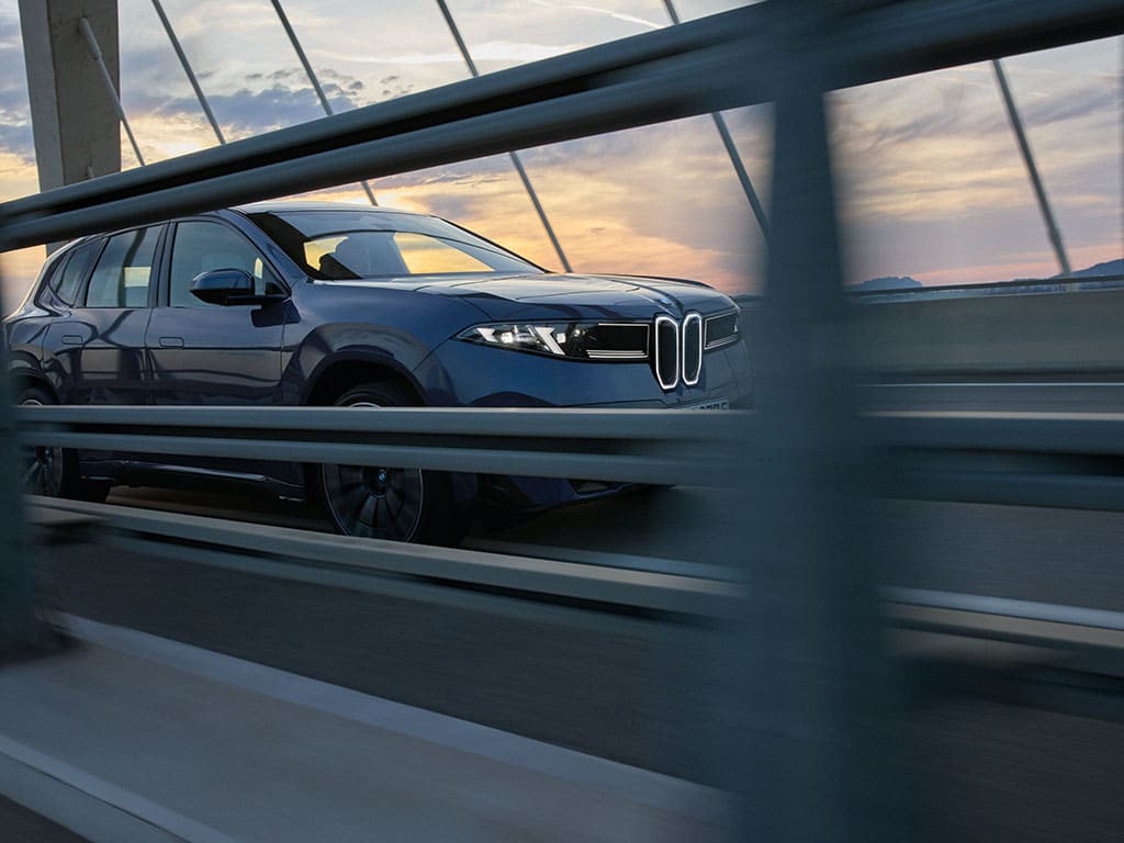 BMW iX3 in Blau fährt über eine moderne Brücke bei Sonnenuntergang, dynamische Seitenansicht mit beleuchteter BMW Niere und urbaner Architektur im Hintergrund.