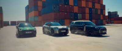 Drei MINI Modelle fahren nebeneinander vor gestapelten Containern auf einem Industriegelände und wirbeln Staub auf.