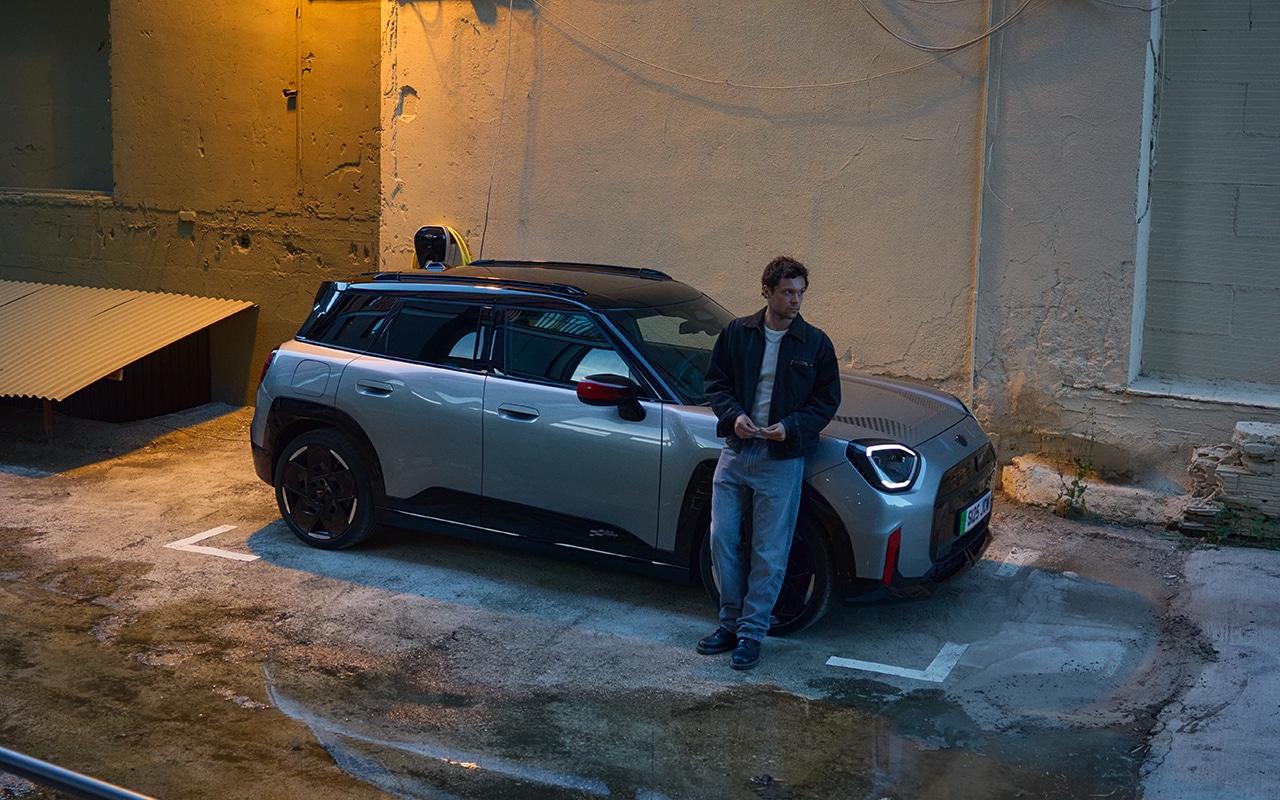 MINI Aceman parkt in einer urbanen Hinterhofszene bei Abendlicht, während ein Mann lässig am Fahrzeug lehnt