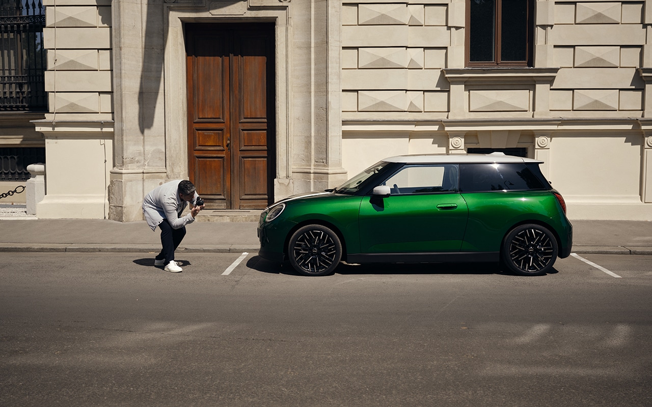 Grüner MINI Cooper parkt am Straßenrand vor einem historischen Gebäude, während ein Fotograf das Fahrzeug aus niedriger Perspektive aufnimmt.